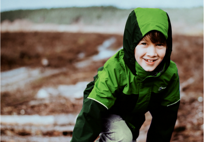 Smilende dreng i grønt og sort overtøj og cowboybukser sidder på hug i i naturen.