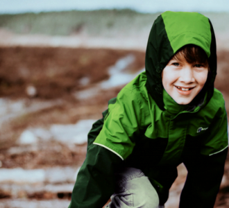 Smilende dreng i grønt og sort overtøj og cowboybukser sidder på hug i i naturen.