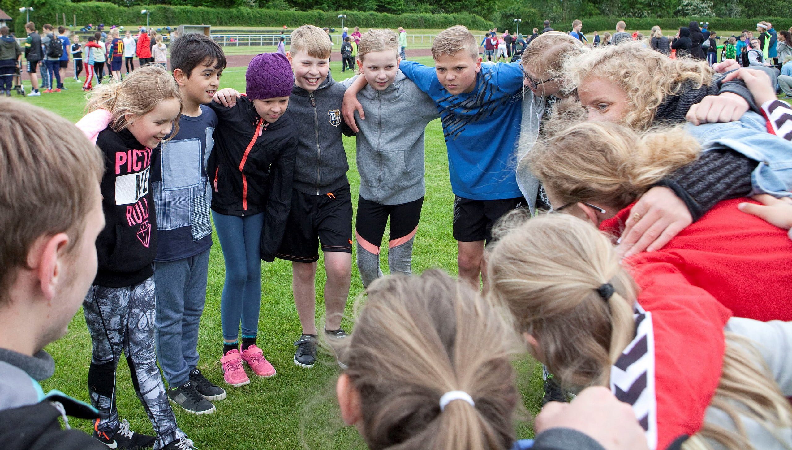 Børn og voksne står i rundkreds og holder hinanden om skuldrene til idrætsarrangement