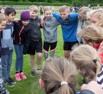 Børn og voksne står i rundkreds og holder hinanden om skuldrene til idrætsarrangement