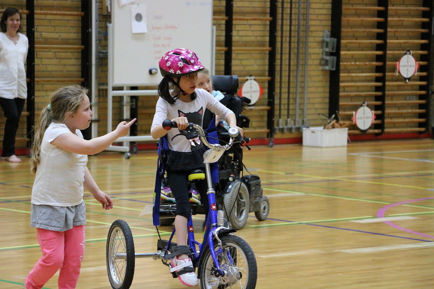 Tre piger i gymnastiksal. Pigen til venstre går rundt. Pigen til højre kører på trehjulet cykel. Pigen i baggrunden kører i el-kørestol.