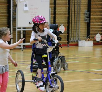 Tre piger i gymnastiksal. Pigen til venstre går rundt. Pigen til højre kører på trehjulet cykel. Pigen i baggrunden kører i el-kørestol.