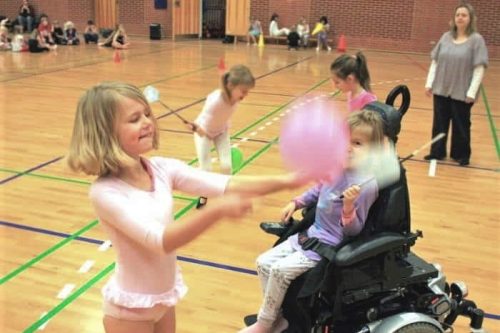 Børn leger med balloner og fluesmækkere i gymnastiksal. Et af børnene sidder el-kørestol.